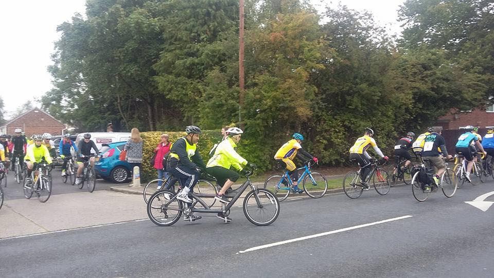 A groupf of men and women cycling along a road, one bike is a tandem with a pilot.